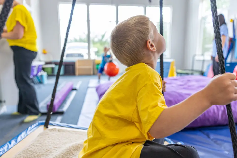 A young boy in a yellow shirt on a swing in the physical therapy gym