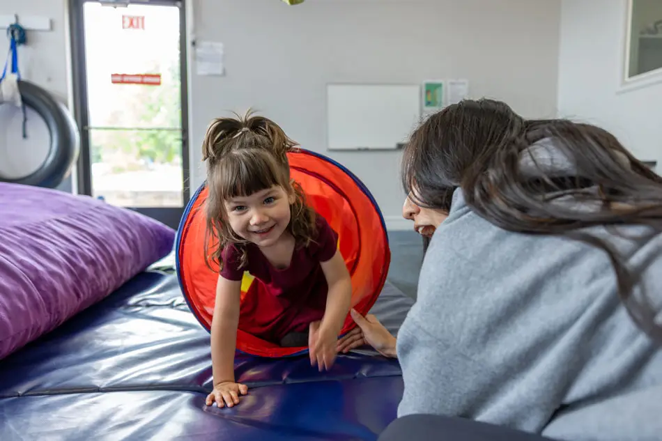 A child crawling through a play tunnel toward a crash pad in the therapy playroom